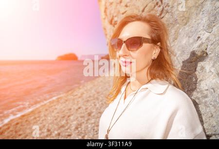 Woman beach sunglasses leaning against rock wall during sunset at seaside coast enjoying vacation Stock Photo
