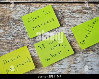 Sticky notes on a wooden table show messages about being open for business and reminders on down payment and investment timing. The notes express urge Stock Photo