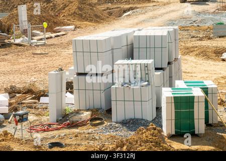 Stacked insulated concrete blocks secured with green straps on construction site. Surrounding environment shows raw earth, gravel, and additional materials waiting to be used. Highlights efficient material organization, construction readiness, project planning, and logistical management in building operations. Suitable for architectural presentations, construction industry reports, real estate marketing materials, and educational content on building processes. Stock Photo