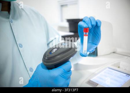 A laboratory technician wearing blue nitrile gloves scans a test tube with a handheld barcode scanner, focusing on sample tracking and diagnostic test Stock Photo