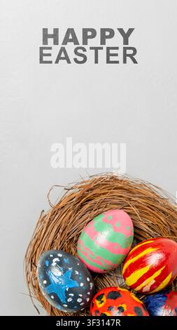 A vertical greeting card for Easter featuring hand-painted colorful eggs resting in a rustic nest made of twigs, set against a clean gray background w Stock Photo