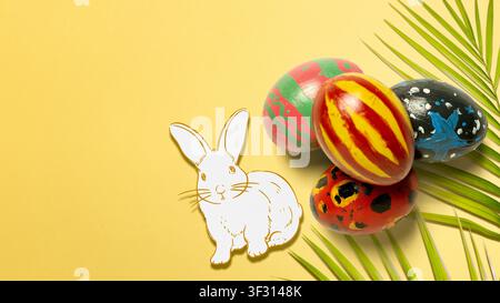 A festive arrangement featuring four hand-painted Easter eggs, a white bunny drawing, and a green palm leaf on a solid yellow background with ample co Stock Photo