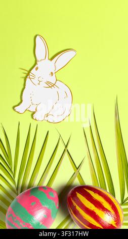 A creative Easter layout on a lime green background featuring a hand-drawn white rabbit, two vibrantly painted eggs, and decorative palm fronds Stock Photo