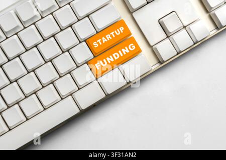 High-angle shot of a white computer keyboard featuring two prominent orange keys labeled Startup and Funding, symbolizing business investment and entr Stock Photo