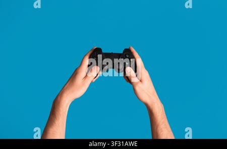 Young man holding videogame joystick on blue background, closeup of hands Stock Photo