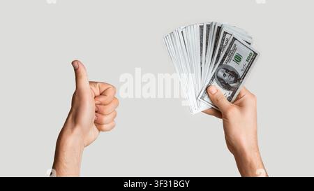 Close up of successful man holding fan of money and showing thumb up gesture on light grey background, panorama Stock Photo