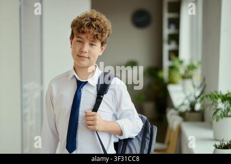 Portrait of Caucasian teenager boy carrying backpack and wearing school uniform, looking at camera in hallway. Ideal for education marketing, back to school campaigns, student services Stock Photo