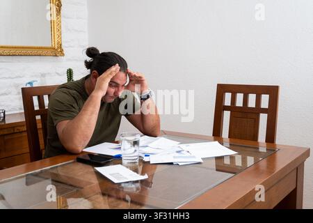 Young adult man sitting at a table with papers, feeling worried and stressed about bills Stock Photo