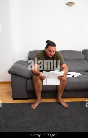 Young adult man sitting on sofa receiving distressing debt notice Stock Photo