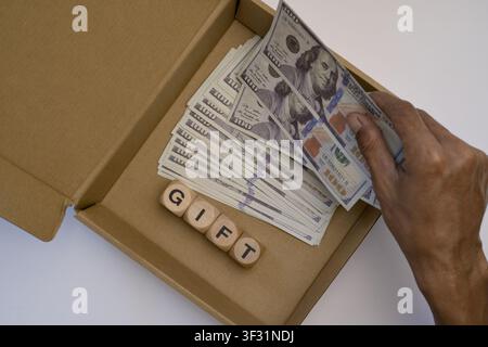 A stack of US dollar bills and wooden blocks spelling gift inside an open cardboard box. Stock Photo