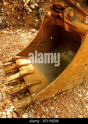 Front view of a large, heavy excavator steel bucket with sharp teeth resting on a rocky ground at an active construction or mining site. Stock Photo