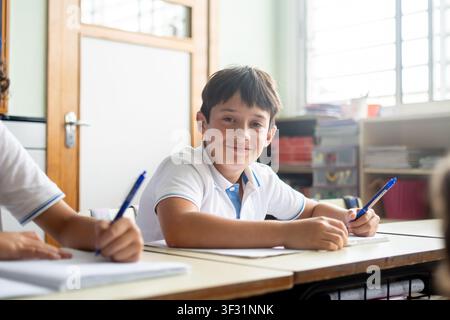 Smiling elementary school boy looking at the camera while writing in his notebook during class. Stock Photo