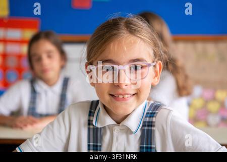 Portrait of a smiling elementary school girl wearing glasses and school uniform while looking at the camera in a bright classroom. Stock Photo