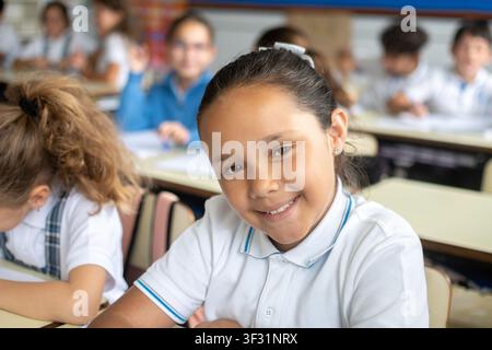 Happy elementary school girl smiling at the camera while sitting at her desk during class. Stock Photo