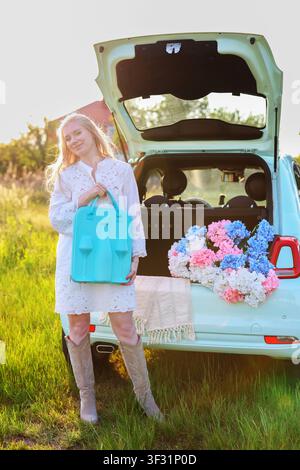 Smiling young woman holding a turquoise picnic case beside a flower-filled car trunk at golden hour in a peaceful countryside setting Stock Photo