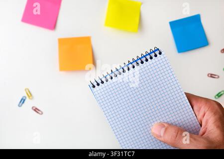 Close-up of a hand holding a blank graph-paper notebook over a desk with colorful sticky notes and paper clips, representing planning, organization, a Stock Photo