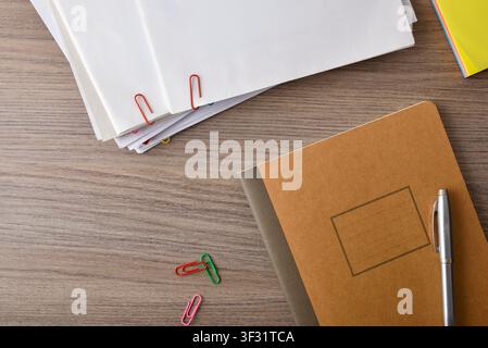 Top view of a notebook, stacked papers, paper clips and pen on a wooden desk with copy space, illustrating organization, office work and productivity. Stock Photo