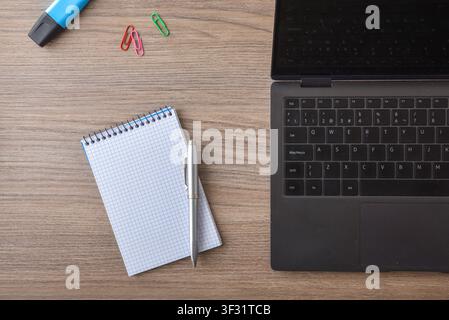 Top view of a wooden office desk with a laptop, spiral notebook, pen, paper clips, and marker. Clean workspace concept with copy space. Stock Photo