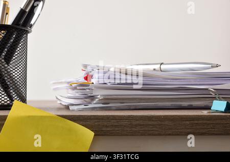 Side view of a pile of office documents with clips and a silver pen resting on top, beside a mesh pen holder on a wooden desk. Stock Photo