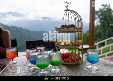 An afternoon refreshment table with layered fruit, pastries and colorful drinks on glass goblets arranged beside mountain view, inviting relaxed outdo Stock Photo