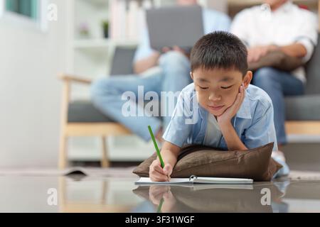 Asian boy doing homework on floor with parents working in background Stock Photo