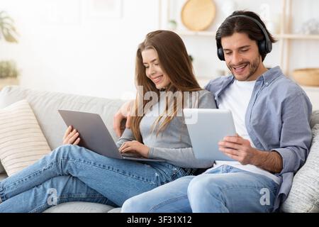 Smiling man and woman using electronic devices, home interior Stock Photo