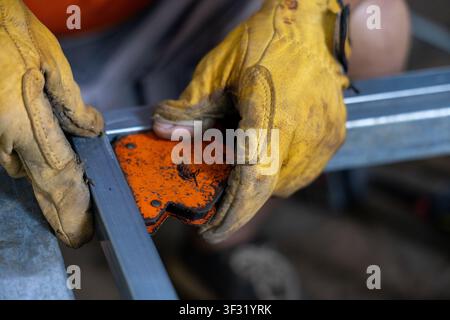 A gloved hands hold orange metal corner clamp while aligning rectangular steel tubing for welding, showing textured leather gloves, worn paint and foc Stock Photo