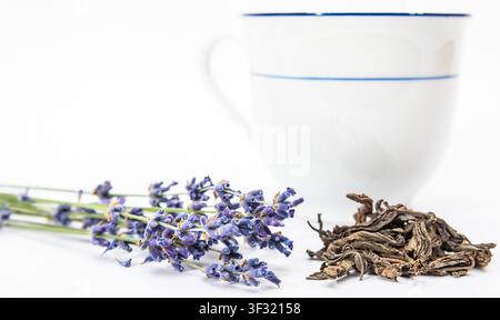 Lavender flowers are placed beside dried leaves next to a white cup on a plain white surface. Stock Photo