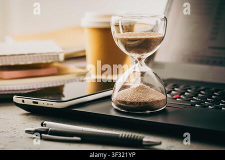 Hourglass on a laptop with office supplies and coffee, symbolizing time management Stock Photo