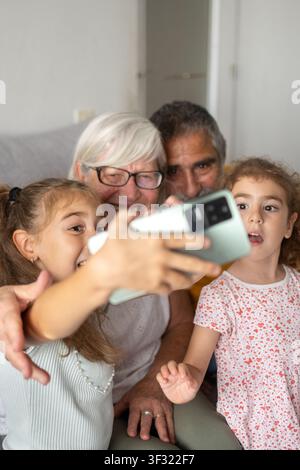 Happy grandparents taking a selfie with their granddaughters using a smartphone while spending quality time together at home. Stock Photo