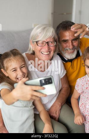 Happy grandparents taking a selfie with their granddaughters using a smartphone while spending quality time together at home. Stock Photo