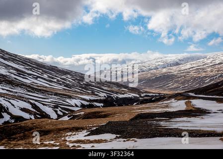 A vast panorama shows a gorge between Icelandic mountains covered in snow streaks and golden fields. On the right side, a lonely asphalt road winds un Stock Photo