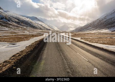 A lonely asphalt road stretches through a scenic valley between snow-covered mountains in Iceland. The afternoon sun breaks through clouds, casting br Stock Photo