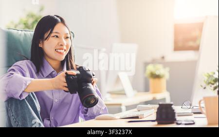 Happy Asian Female Photographer Smiling While Preparing Camera In Modern Office Stock Photo