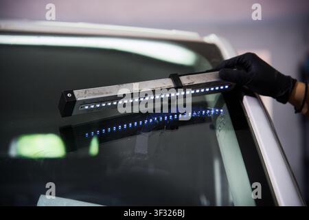 Automotive technician holds an ultraviolet lamp over damaged car glass to cure the resin during a windshield repair process. Stock Photo