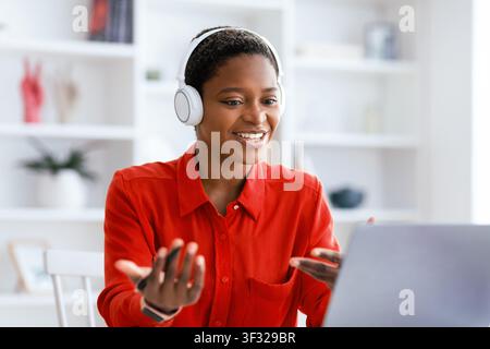 Woman in Red Shirt Using a Laptop With Headphones During an Online Meeting in Bright Room Stock Photo