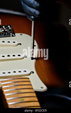 Close-up shot of a guitar technician or luthier using a screwdriver to fasten the pickguard on a sunburst electric guitar. Professional instrument maintenance, setup, electronics installation, and luthiery craft in a dark workshop setting. Ideal for music service, guitar repair shop, craftsmanship, and rock music concepts. Stock Photo