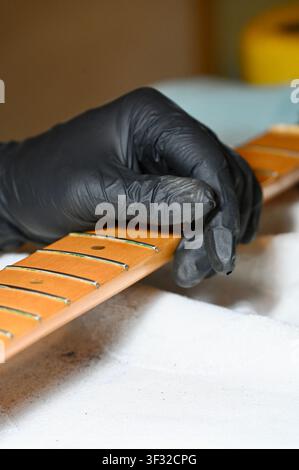Close-up shot of a guitar technician or luthier wearing a black nitrile glove holding a detached electric guitar maple neck with frets during cleaning, polishing, and setup work. Professional instrument maintenance, luthiery craftsmanship, and workshop process. Ideal for music service, repair shop, guitar care, and craftsmanship concepts. Stock Photo