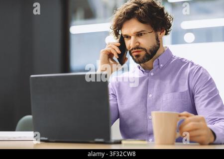 Man on Phone While Working on Laptop in Office Setting During Daytime Hours Stock Photo