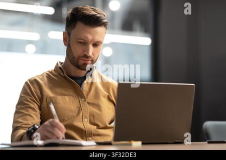 Focused Caucasian Male Writing Notes At Laptop In Modern Office, Business Concept Stock Photo