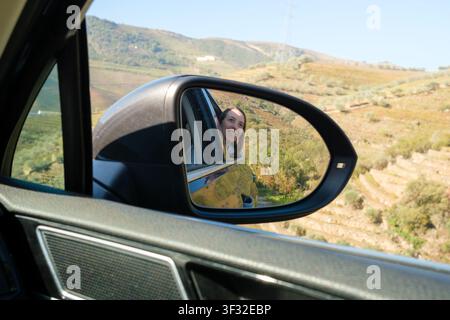 Woman smiling through car window as seen in side mirror during a scenic road trip Stock Photo