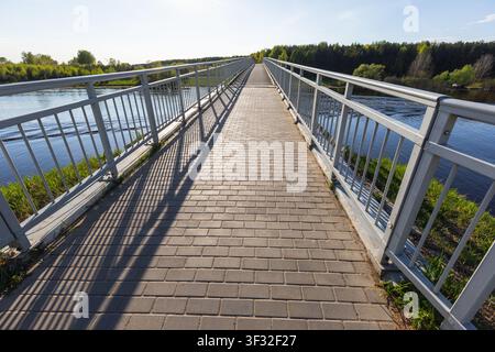 Long pedestrian footbridge over a calm river, showing leading lines, metal railings, brick walkway and strong shadows; a peaceful outdoor scene with t Stock Photo