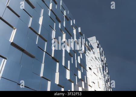 A modern mirrored glass facade composed of repeating geometric panels reflecting the blue sky and sunlight, creating an abstract, rhythmic pattern tha Stock Photo