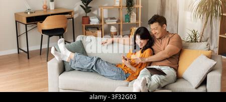Happy Asian Couple Relaxing On Couch, Using Smartphone In Cozy Living Room Stock Photo