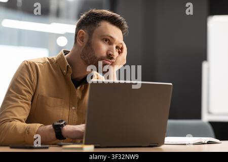 Worried Young Caucasian Man Working On Laptop At Office Desk, Business Concept Stock Photo