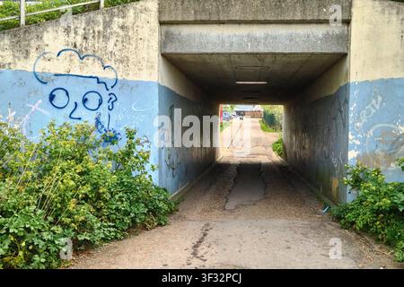 A concrete pedestrian underpass decorated with graffiti tags and spray-painted street art leading toward a distant road on a sunny day. Stock Photo