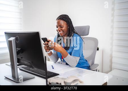 Smiling Black nurse using smartphone in office Stock Photo