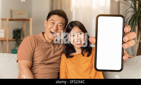 Happy Millennial Asian Couple Enjoying Time Together, Holding Smartphone With Blank Screen, Home Stock Photo