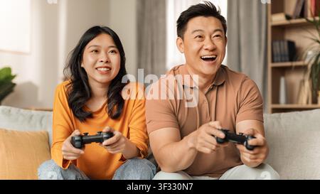 Happy Millennial Asian Couple Playing Video Games In Living Room, Indoor Fun Activity Stock Photo