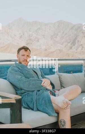 Man in bathrobe sitting on yacht deck with mountains in background Stock Photo